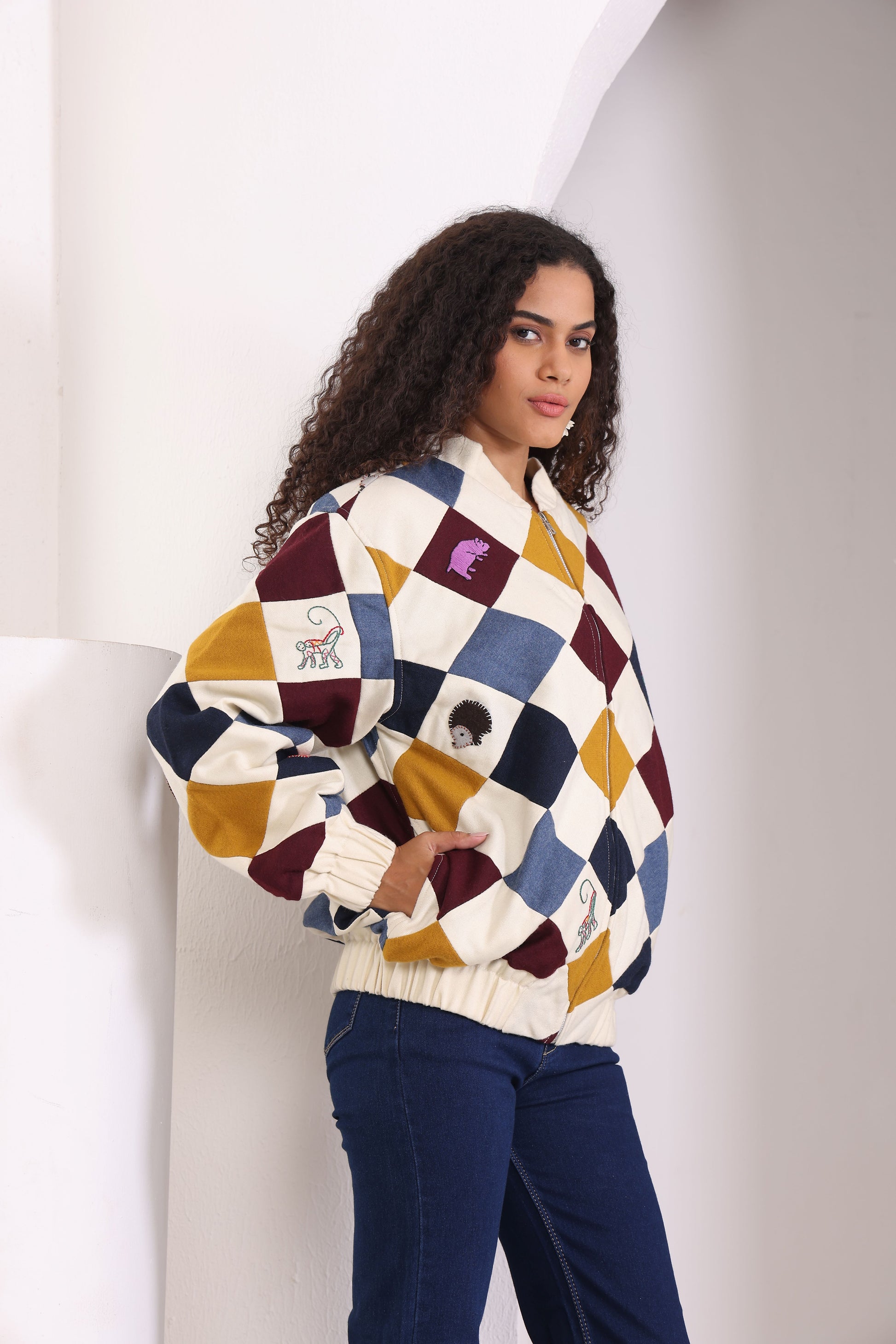 A model wearing a multicolored patchwork bomber jacket with geometric patterns, featuring a pinboard on the left side.