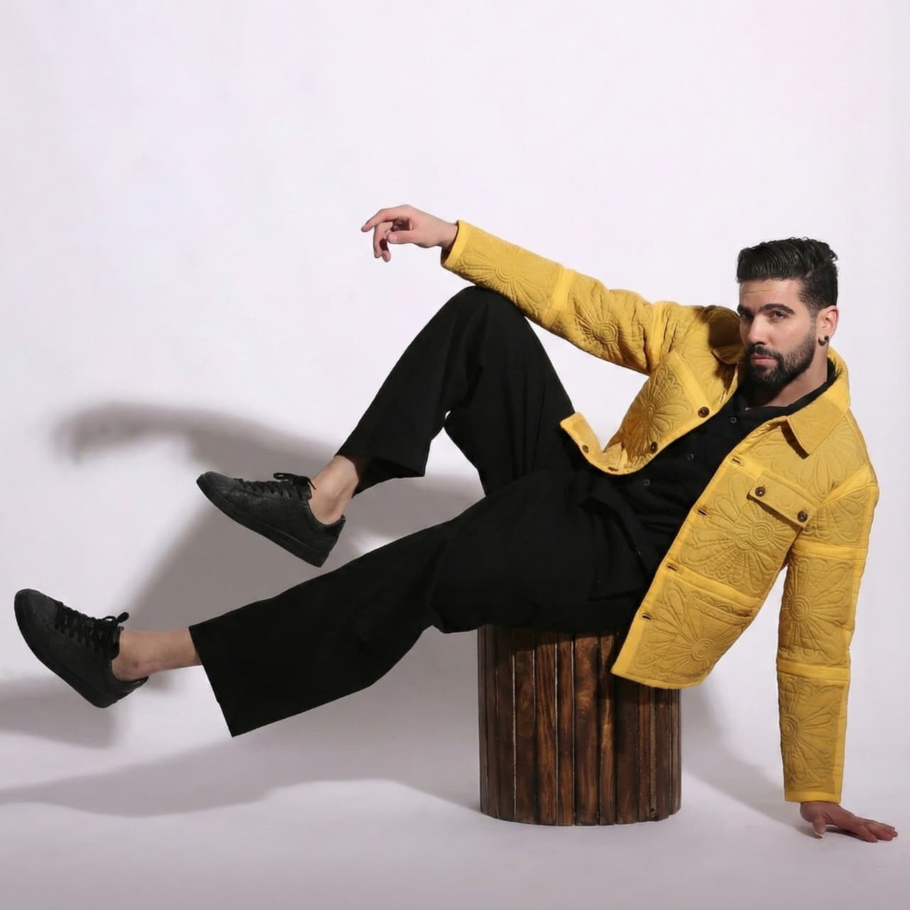 Man in a yellow jacket and black pants sitting on a wooden stool against a white background
